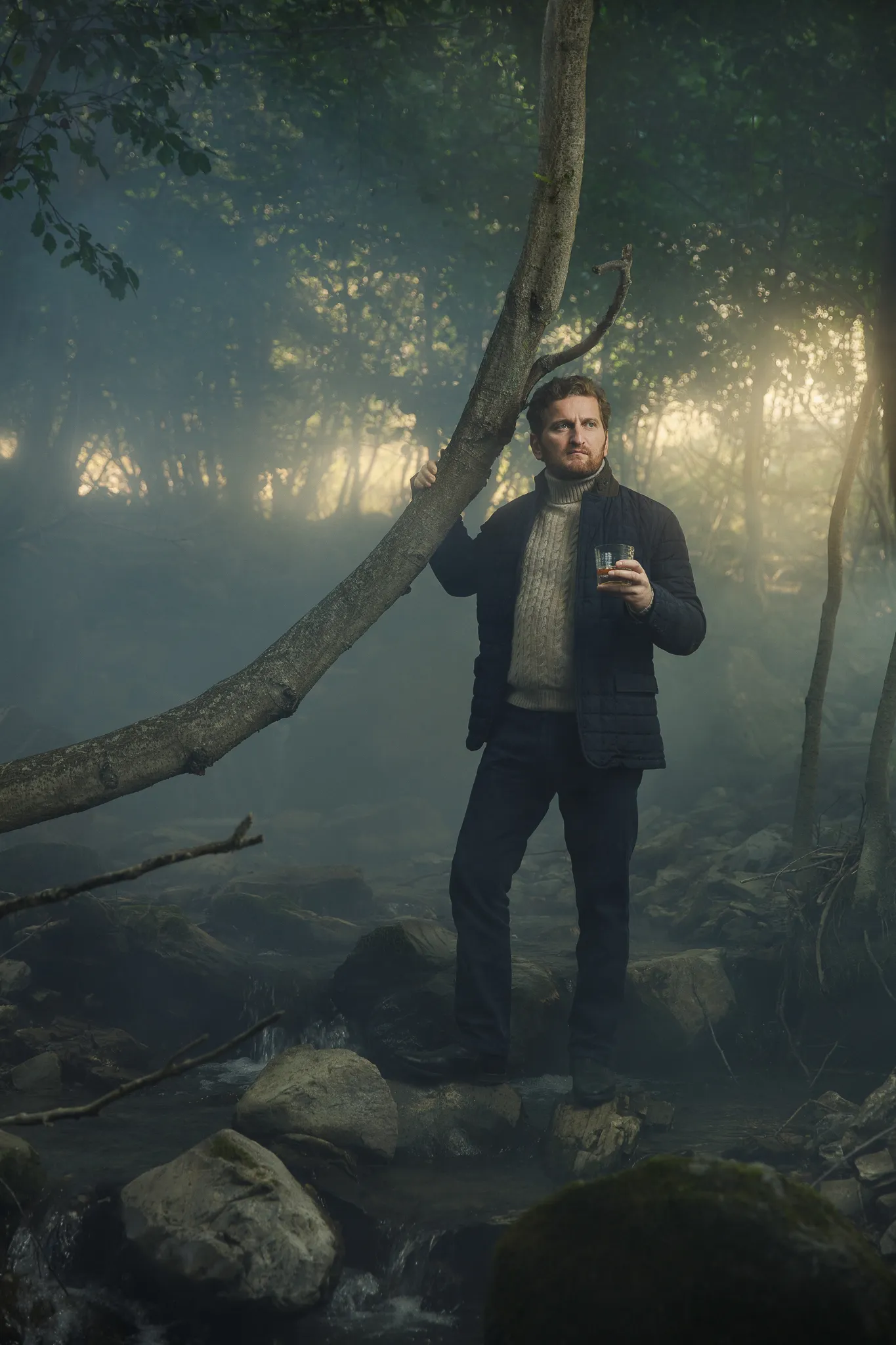 A man in a quilted jacket holds a glass by a stream in a misty forest.
